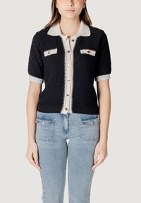 Cardigan nero lavorato a maglia con maniche corte, colletto bianco, tasche anteriori e dettagli con bottoni metallici. Abbinato a jeans azzurri chiari.