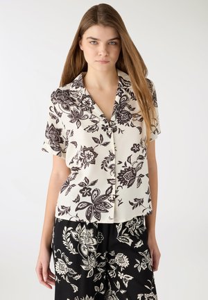 Giovane donna con capelli lunghi che indossa una camicetta floreale a maniche corte abbottonata e pantaloni floreali coordinati su uno sfondo semplice.