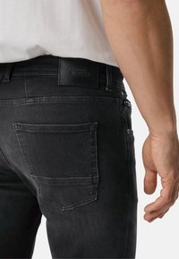 Black denim jeans shown from the back, featuring a single back pocket, a leather tag at the waistband, and a fitted silhouette.