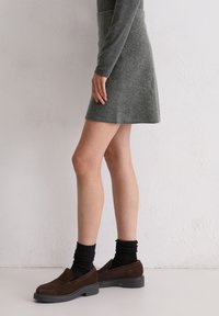 Personne portant une robe à manches longues grise, des chaussettes côtelées noires et des mocassins en daim marron, debout contre un mur blanc texturé.