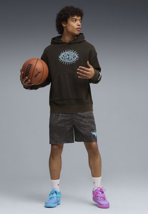 Junger Mann, der einen Basketball hält, trägt einen dunklen Hoodie, gemusterte Shorts, weiße Socken und unterschiedlich farbige blaue und pinke Turnschuhe, steht vor einem grauen Hintergrund.