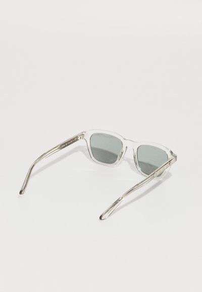 Lunettes de soleil à monture transparente avec des verres gris, présentant des branches fines et texturées avec un motif subtil sur les côtés.