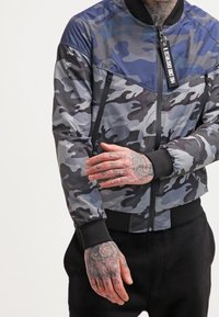 Kurtka bomber w kamuflażu w odcieniach szarości i niebieskiego, z zapięciem na zamek, ściągaczami na rękawach oraz boczną kieszenią z metką.