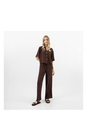 Femme debout portant une chemise marron ample boutonnée et un pantalon large assorti, ainsi que des sandales noires avec des détails en chaîne.