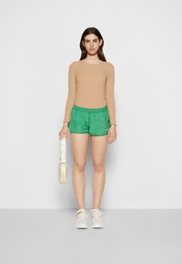 Calções verdes com brilho, cintura elástica, combinados com uma blusa de manga longa bege. A modelo segura uma carteira creme e usa ténis brancos.