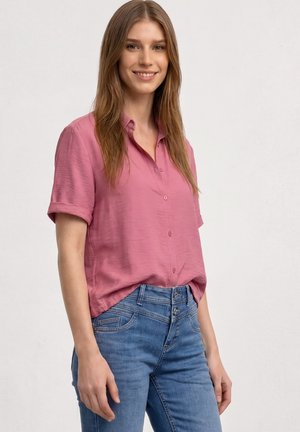 Ung kvinde med langt brunt hår iført en lyserød kortærmet skjorte og blå jeans smilende mod en ensfarvet lys baggrund.