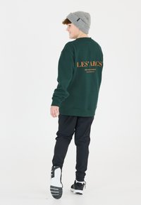 Mörkgrön sweatshirt med "LES' ARCS" tryckt i orange på ryggen. Kombination med svarta byxor och svart-vita sportskor. Grå mössa bärs.