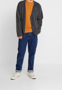 Cárdigan de un gris oscuro y suave, con un corte relajado, superpuesto a una sudadera naranja. Llevado con jeans azules y zapatillas blancas.