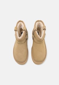 Paire de bottines en daim beige avec doublure en polaire blanche moelleuse et sangles à boucle latérale, vue de dessus sur fond blanc.