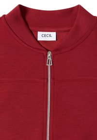 Veste rouge zippée avec tirette argentée et étiquette blanche portant la mention "CECIL" au niveau de l'encolure.