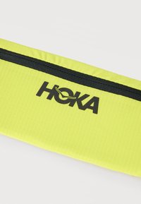 Κίτρινο υφασμάτινο τσαντάκι με μαύρο φερμουάρ και το λογότυπο HOKA τυπωμένο με μαύρο στο μπροστινό μέρος.