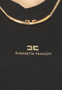 T-shirt nera con una superficie liscia, con logo dorato "ELISABETTA FRANCHI" e un'accenno di collana dorata curvata sul collo.