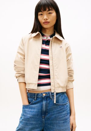 Noor naine sirgete tumedate juustega, kes kannab beeži tõmblukuga jope, triibulist mereväe, valge ja punasega polosärki ning siniseid kõrge vöökohaga teksaseid, käed taskutes.