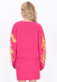 Jaskrawo różowy sweter z żółtymi wzorami kwiatowymi na obu rękawach. Miękka tekstura, dzianinowy materiał, prążkowany dół i mankiety oraz luźny krój.