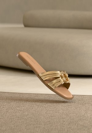 Sandale plate beige avec des lanières métalliques dorées et une boucle décorative en or, flottant au-dessus d'un tapis texturé avec en arrière-plan un canapé beige.
