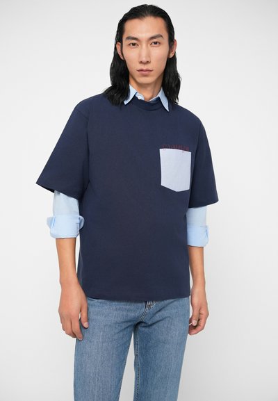 Bărbat cu păr lung negru, purtând o cămașă button-up ușoară, albastră, stratificată și o cămașă scurtă, bleumarin, cu buzunar pe piept de culoare albastră deschisă, stând în picioare.