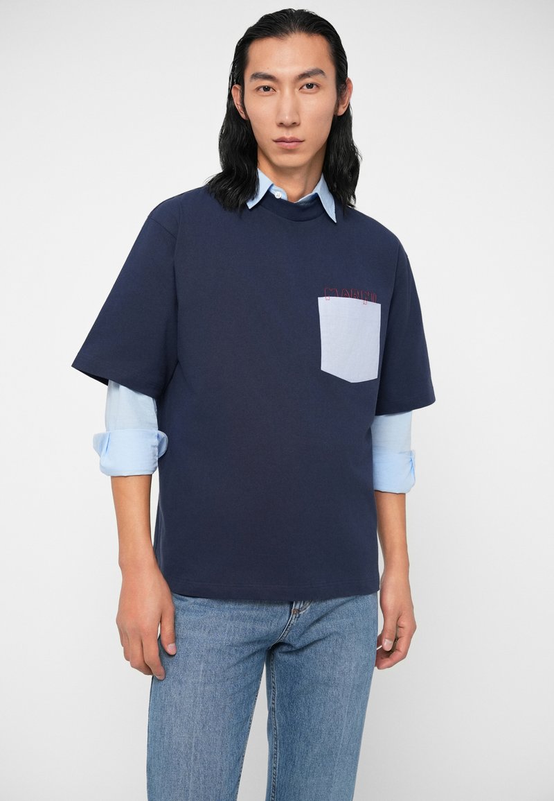 Mann mit langen schwarzen Haaren, der ein gestuftes hellblaues Hemd und ein dunkelblaues Kurzarmhemd mit einer hellblauen Brusttasche trägt, stehend.