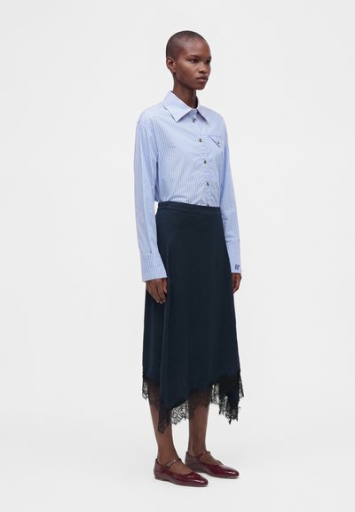 Femme portant une chemise à rayures bleu clair boutonnée et une jupe midi noire avec un ourlet en dentelle, assortie à des chaussures plates rouge foncé, debout de profil.