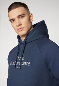 Sweat à capuche bleu marine en tissu doux avec un logo "Peak Performance" brodé en argent à l'avant et des cordons ajustables au niveau de la capuche.