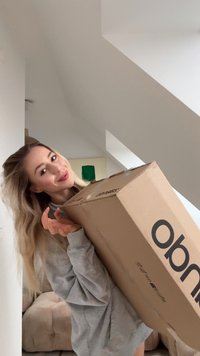 Kartónová krabica s vytlačeným logom, držená pred svetlou stenou. Krabica je obdĺžnikového tvaru s viditeľnou páskou na vrchu.