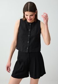 Femme portant un haut noir sans manches boutonné et un short plissé noir taille haute, regardant vers le bas avec un léger sourire sur un fond uni.