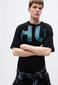 Μαύρο κοντομάνικο μπλουζάκι με μεγάλο πράσινο-μπλε γράμμα "HU" στο μπροστινό μέρος. Συνδυάζεται με γκρι καρό πουκάμισο δεμένο γύρω από τη μέση.