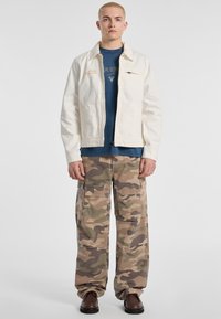 White denim jacket with front zipper, blue T-shirt with logo, and camo cargo pants featuring large pockets, worn with dark brown shoes.