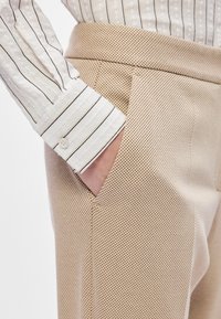 Calças bege, texturizadas, com um padrão em rede e um bolso lateral, usadas com uma camisa branca, riscada, com botões e um leve brilho.