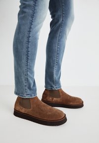 Botas de ante marrón hasta el tobillo con paneles laterales elásticos, suelas de goma negra gruesa, combinadas con jeans ajustados azul claro.