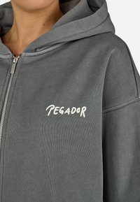 Γκρι φούτερ με φερμουάρ, διαθέτει μεγάλη κουκούλα και μπροστινή τσέπη, με την ένδειξη "TEGADOR" εκτυπωμένη σε λευκό στην αριστερή πλευρά του στήθους. Μαλακή υφή υφάσματος.