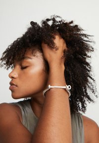 Mujer con los ojos cerrados tocando su cabello rizado, con una blusa sin mangas y una pulsera plateada en la muñeca.