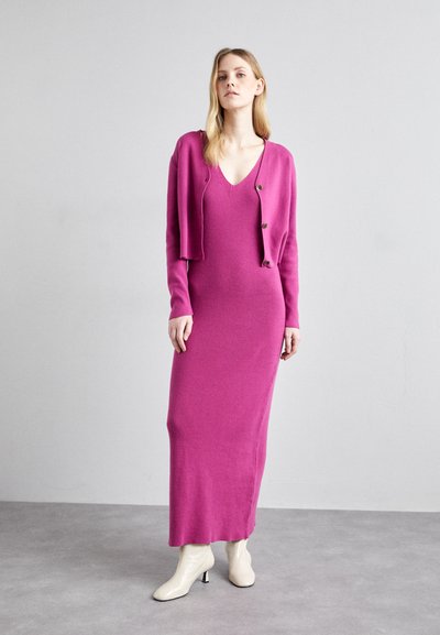 Cardigan tricoté rose avec des boutons et une robe sans manches assortie. La robe ajustée présente un décolleté en V profond et une longueur longue. Bottines blanches à la cheville.