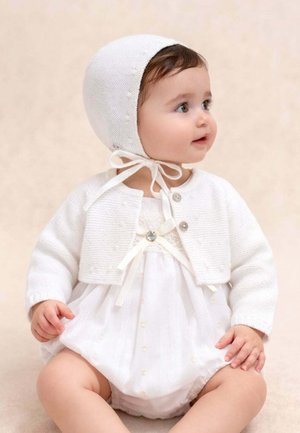 Bébé portant un bonnet en tricot blanc et un cardigan sur une robe blanche, assis et regardant vers la droite sur un fond neutre.