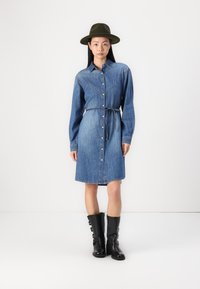 Denim srajčna obleka z gumbi spredaj, dolgimi rokavi in pasom. Nosi se z črnimi usnjenimi škornji do kolen in zeleno klobukom.