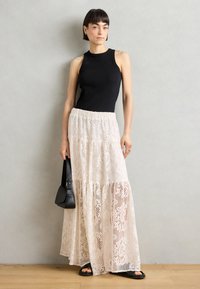 Schwarzes ärmelloses Top kombiniert mit einem cremefarbenen, gestuften Spitzen-Maxikleid. Das Model hält eine kleine schwarze Tasche. Der Boden ist aus Holz, der Hintergrund ist grau.