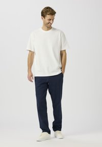 Balta īsroku t-krekls ar apaļu apkakli, saskaņots ar tumši ziliem biksēm un baltiem sporta apaviem, stāvot uz vienkārša fona.