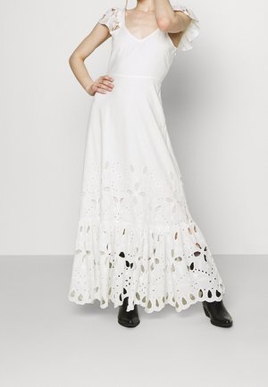 Femme portant une longue robe blanche avec des découpes florales à l'ourlet et des manches courtes, associée à des bottines noires.