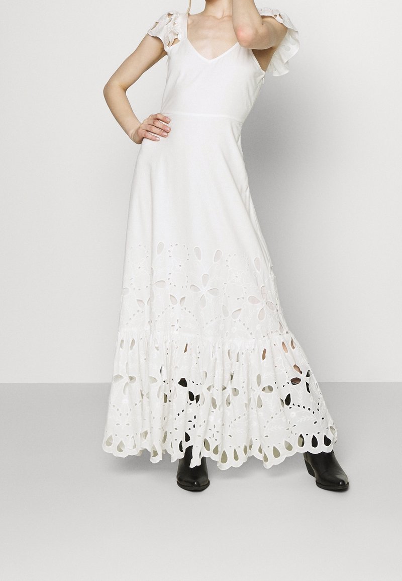 Femme portant une longue robe blanche avec des découpes florales à l'ourlet et des manches courtes, associée à des bottines noires.