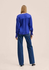Camicia lunga in satin blu con maniche a sbuffo e polsini abbottonati, abbinata a jeans in denim blu a gamba larga, con orlo sfilacciato.