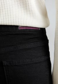 Black denim jeans with visible seam detailing, a purple label at the waist, paired with a textured cream-colored sweater.