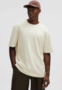 Krémové tričko s krátkym rukávom vyrobené z mäkkého materiálu, s uvoľneným strihom. Nosené s tmavými nohavicami a svetlohnedým klobúkom.