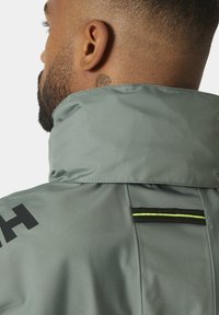 Chaqueta impermeable gris claro con cuello alto, logo negro y una franja de acento negra y amarilla en la parte posterior. Tejido suave y texturizado.