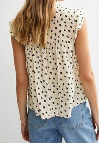 Blusa beige chiaro con pois neri, maniche a sbuffo e vestibilità ampia. Presenta un retro plissettato e un design informale. Abbinata a jeans blu.
