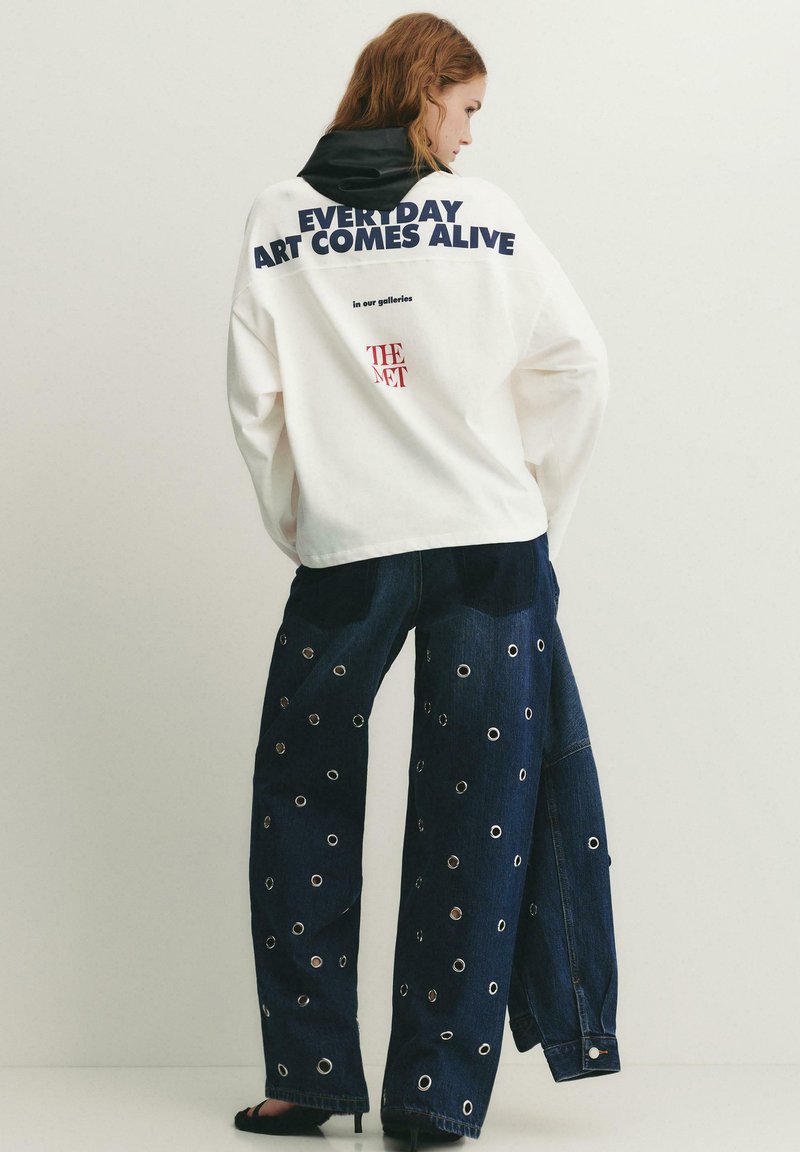Vrouw met een witte trui met de tekst "EVERYDAY ART COMES ALIVE" en donkerblauwe, wijd uitlopende jeans met zilveren oogjes, staand tegen een effen achtergrond.