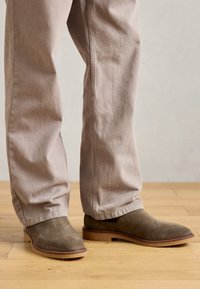 Botas de ante marrón hasta el tobillo con un acabado suave, puntera redonda y suela baja en capas. Combinadas con pantalones de corte relajado en beige claro.