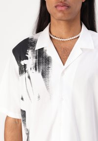 Camicia a maniche corte con bottoni di colore bianco, decorata con una grafica astratta nera su un lato. Presenta un colletto classico e un tessuto liscio e strutturato.