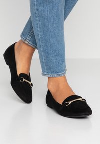 Pieds en jeans bleu clair portant des mocassins en daim noir avec un détail de barre métallique argentée sur un sol blanc.