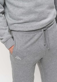 Isik, kes kannab halli paelaga spordipükse ja sobivat halli pusa, käsi vasakus taskus.