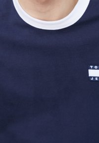 Marineblaues T-Shirt mit einem weißen Rundhalsausschnitt. Verfügt über ein kleines Markenpatch, das zwei weiße horizontale Streifen umfasst. Glatte Textur.