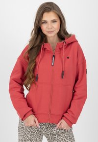 Felpa rossa con zip, realizzata in un tessuto morbido, con cappuccio regolabile, tasca frontale e dettagli neri. Abbinata a pantaloni con stampa leopardata.